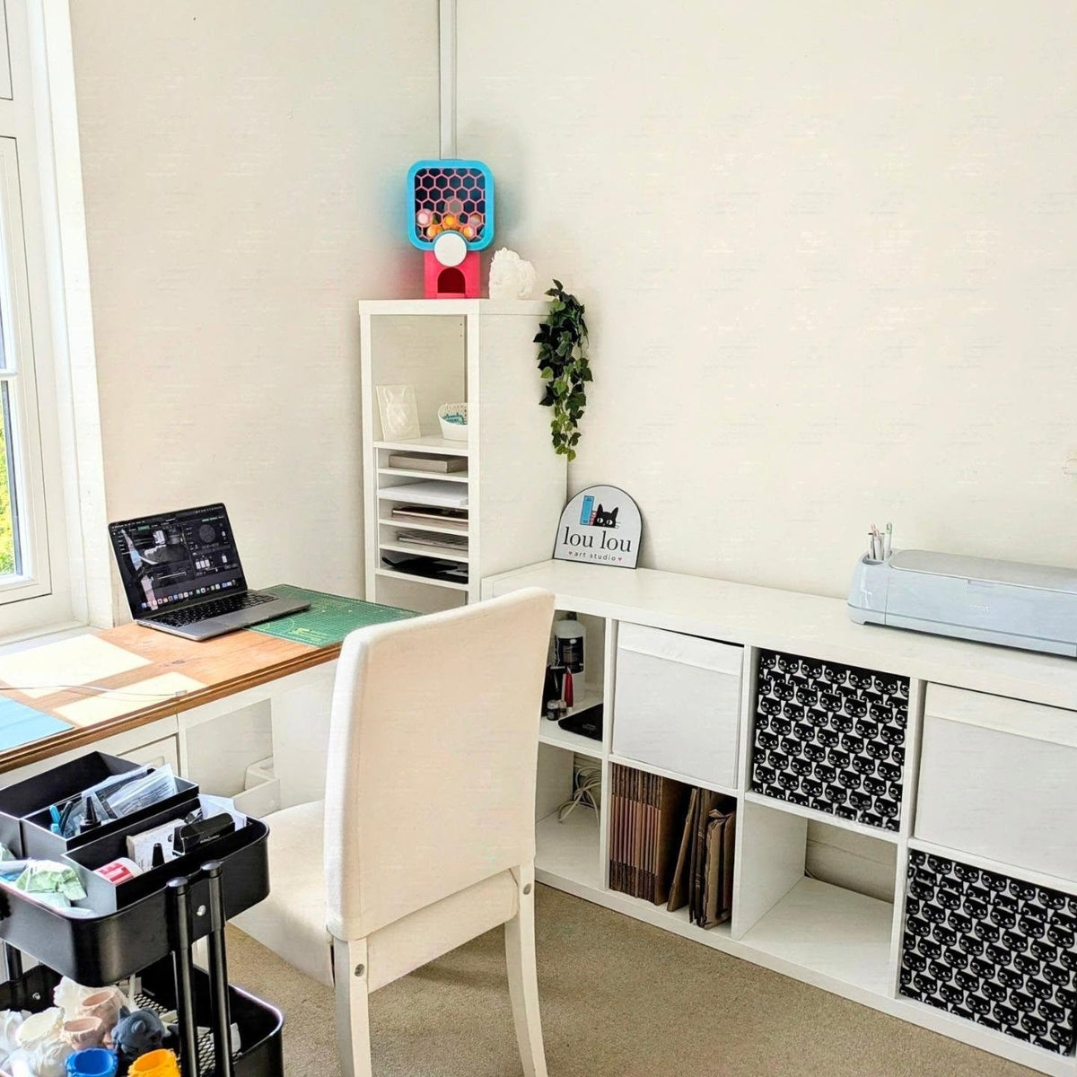 A styled home office corner featuring a white desk, a matching chair, a white shelving unit with storage cubbies and black-and-white patterned bins, and a small gumball machine decoration on top.