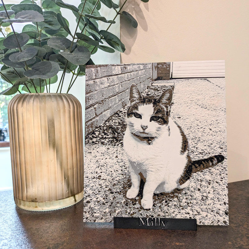 A custom pet portrait canvas print of a white, gray, and black cat named 'Milly' on a black wooden base, styled on a tabletop next to a ribbed glass vase with green leaves.