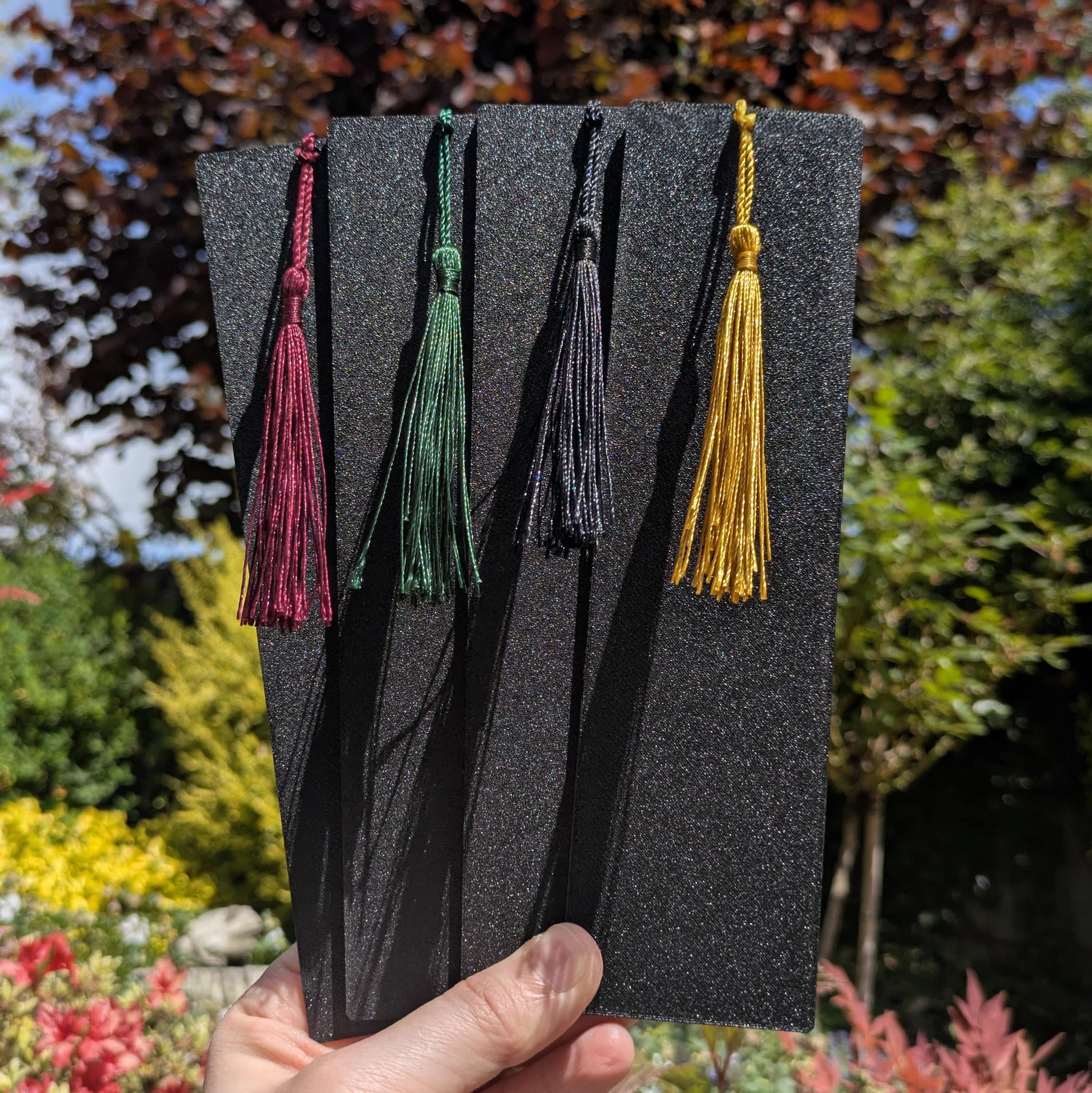 Four velvet tassel bookmarks in burgundy, green, black, and gold, displayed on a black textured card held by a hand against a blurred garden background.